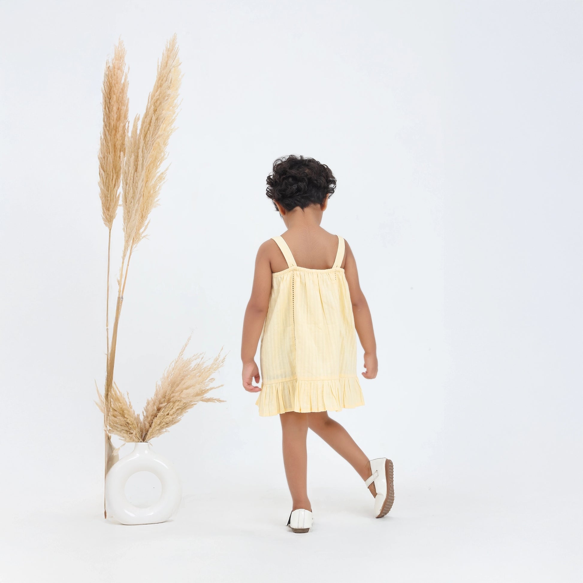 Child in a yellow dress walking away from the camera with dried pampas grass on a white background